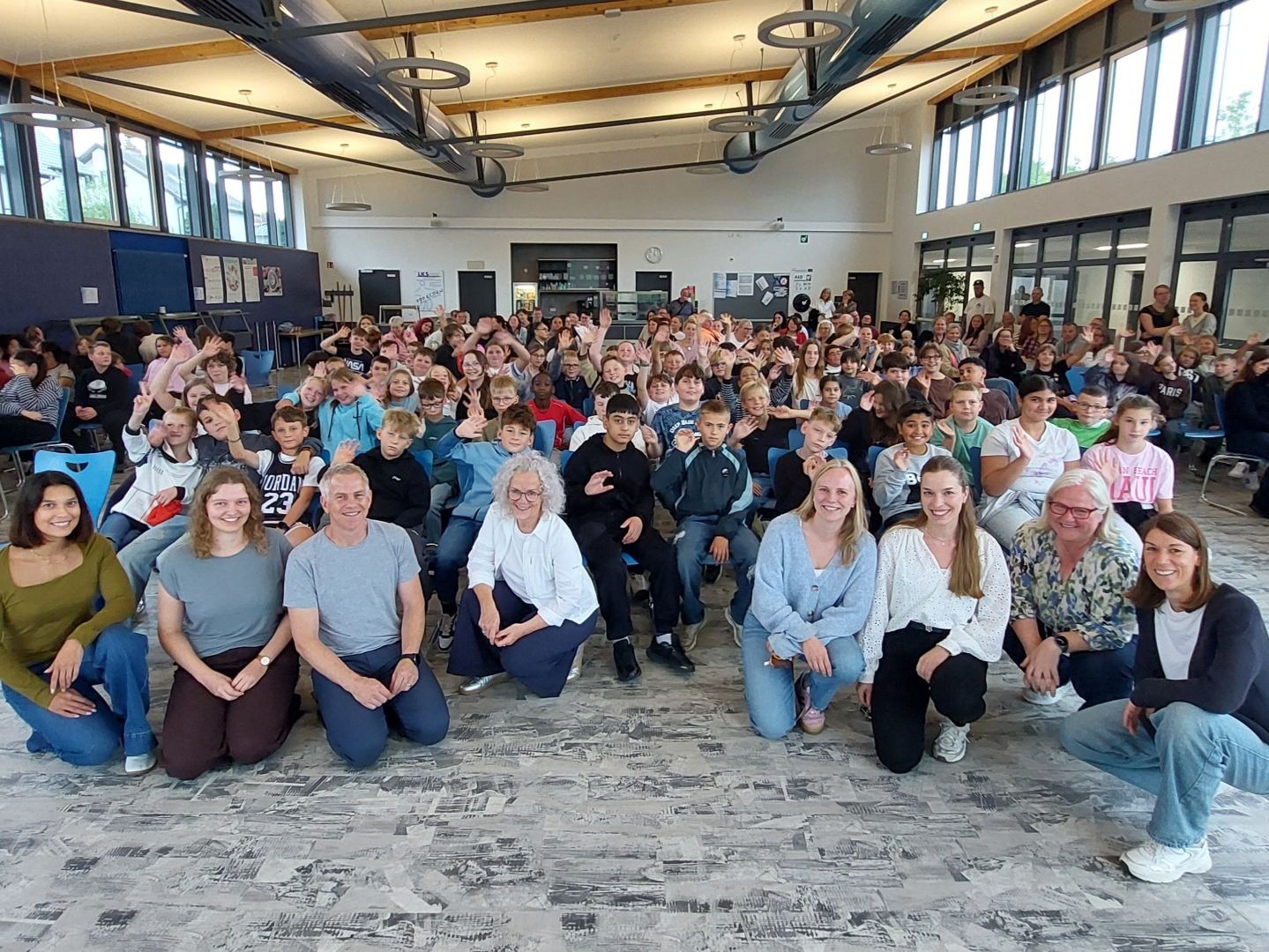 Viele winkende Fünftklässler und lächelnde Erwachsene sitzen in einer großen Schulaula für ein Gruppenfoto