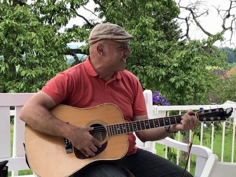 Ennio spielt lächelnd Akustikgitarre im Freien vor grünen Bäumen auf einer weißen Bank