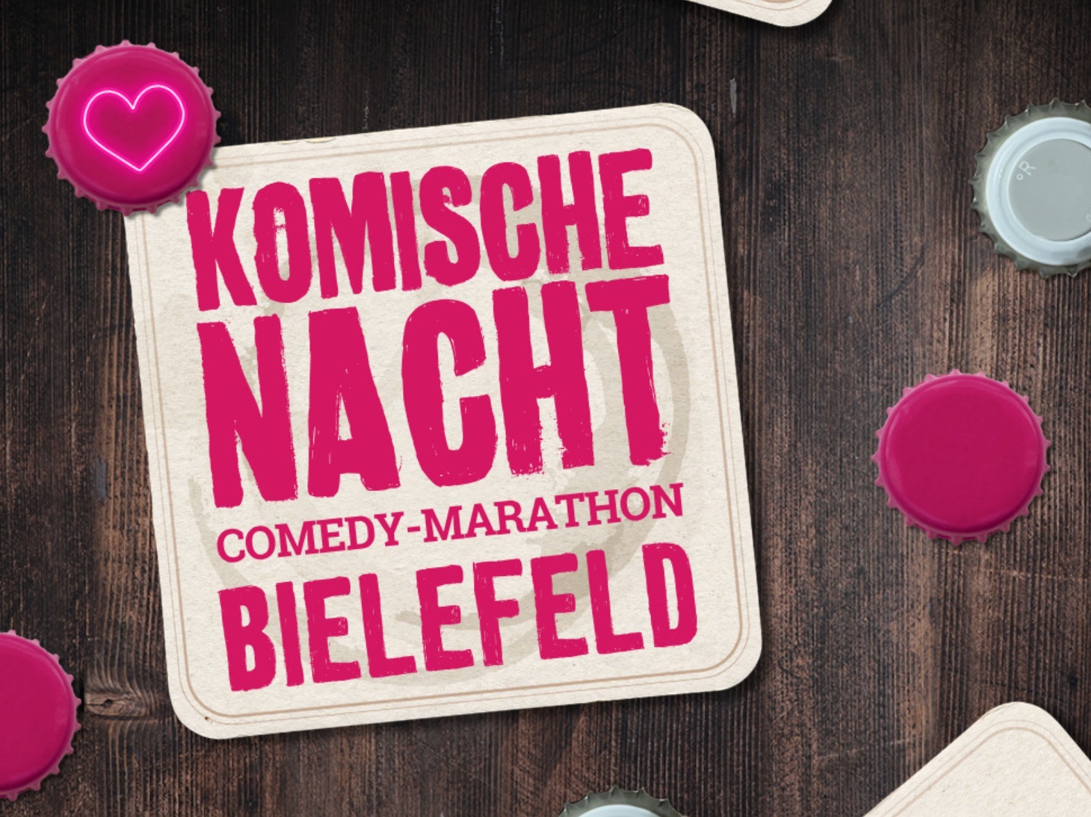 Heller Bierdeckel mit der rosa Aufschrift "Komische Nacht Comedy-Marathon Bielefeld" auf dunklem Holztisch, umgeben von
