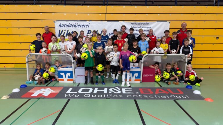 Lächelnde Kinder und Erwachsene in Fußballtrikots posieren mit Bällen in einer Sporthalle vor Bannern