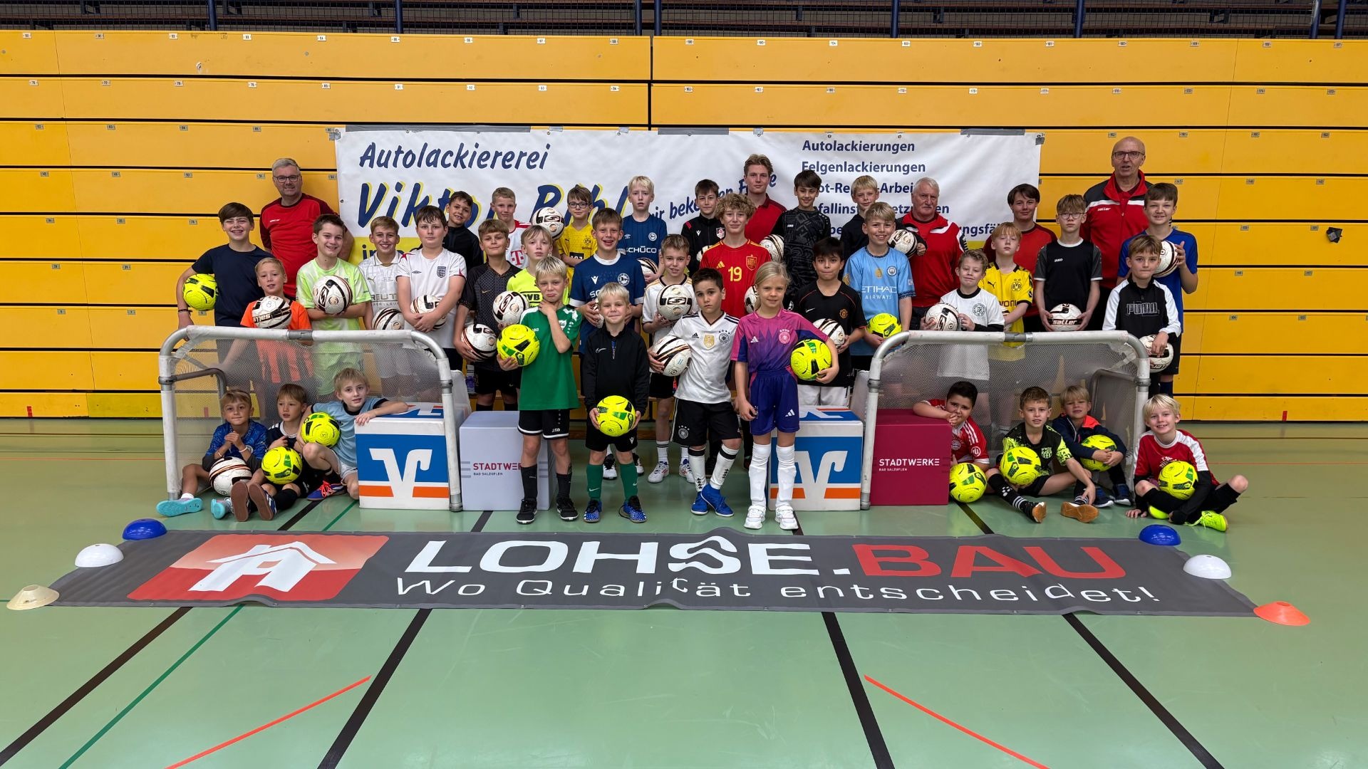 Lächelnde Kinder und Erwachsene in Fußballtrikots posieren mit Bällen in einer Sporthalle vor Bannern