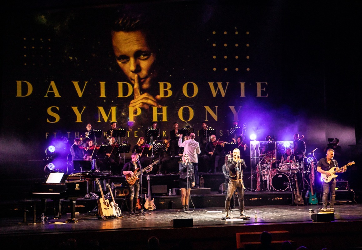 Live-Konzert auf Bühne mit Orchester, Dirigent, Band und Sängern, darüber eine "DAVID BOWIE SYMPHONY"-Leinwand