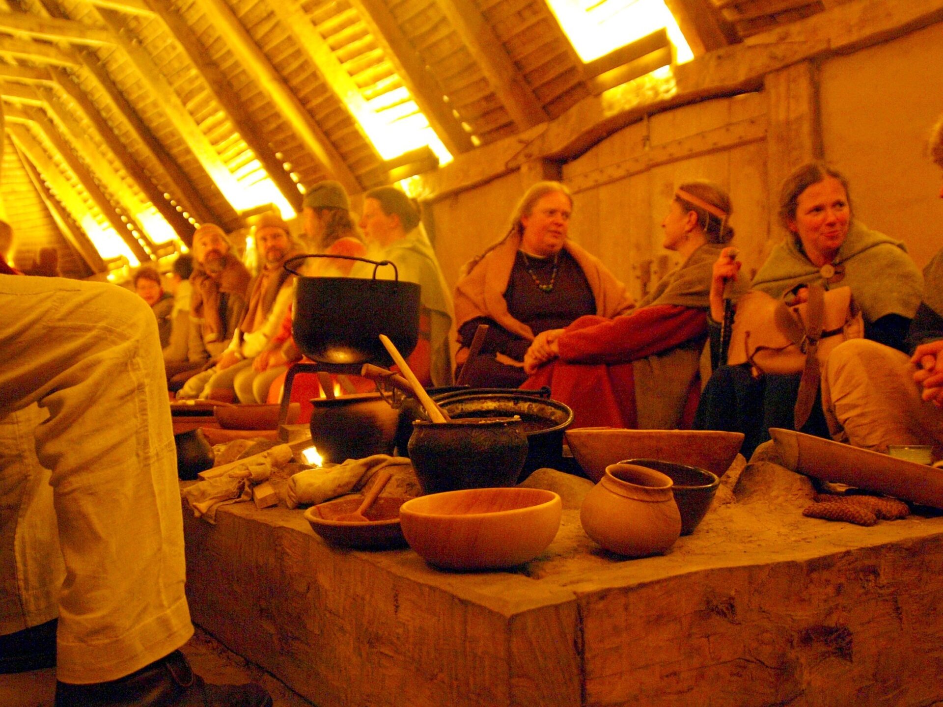 Wikinger sitzen in einem hölzernen Langhaus um brodelnde Töpfe und Holzschüsseln und unterhalten sich