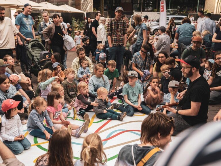 Kinder und Erwachsene sitzen auf einer weißen Matte mit bunten Linien und sehen einem Gitarrenspieler zu
