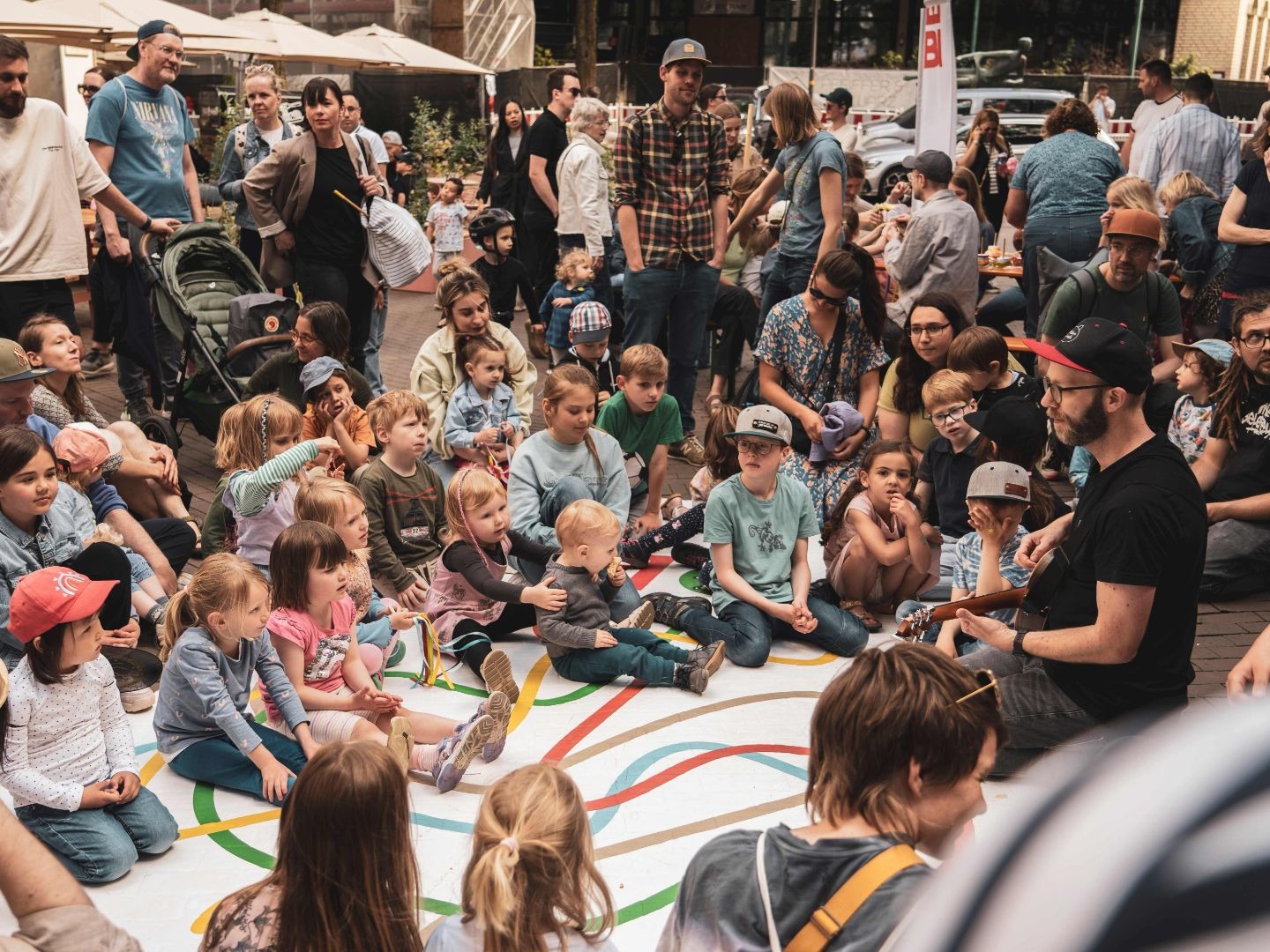 Kinder und Erwachsene sitzen auf einer weißen Matte mit bunten Linien und sehen einem Gitarrenspieler zu