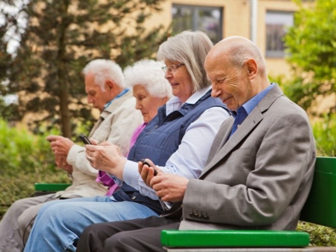 Vier Senioren sitzen draußen auf einer Parkbank und bedienen ihre Smartphones