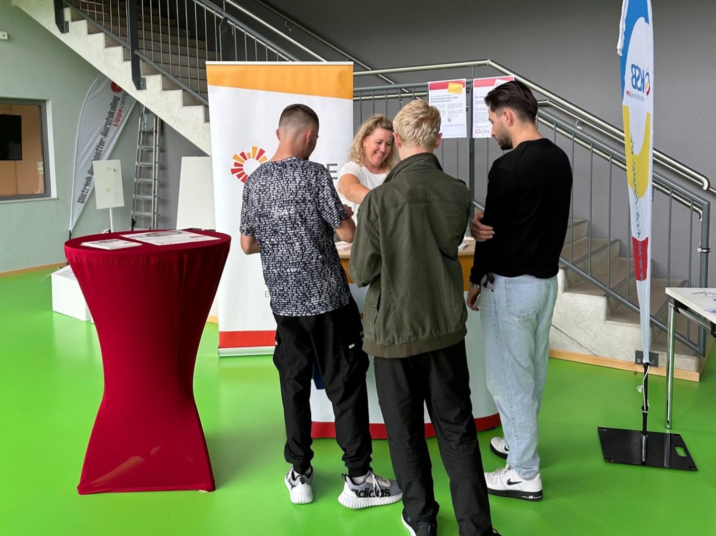 Vier Personen stehen an einem Infostand mit orange-weißem Banner auf grünem Boden in einem Gebäude