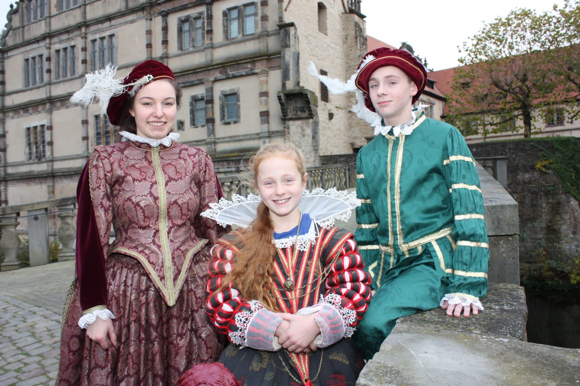 Drei Personen in Renaissance-Kostümen lächeln vor dem Weserrenaissance-Museum an einer Steinmauer