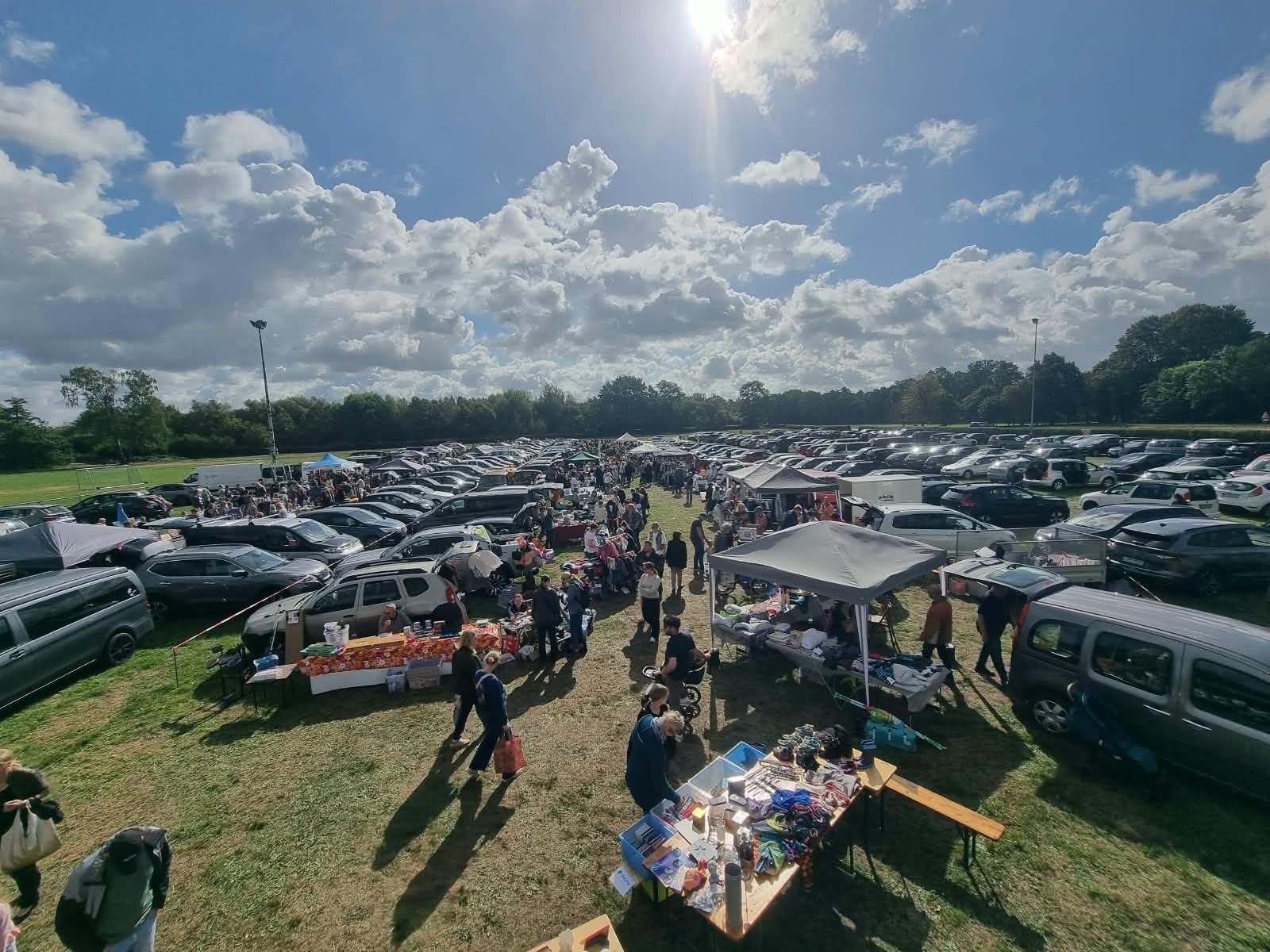 Großer Flohmarkt auf einer Wiese mit vielen Autos, Marktständen und Menschen unter blauem Himmel mit Wolken in