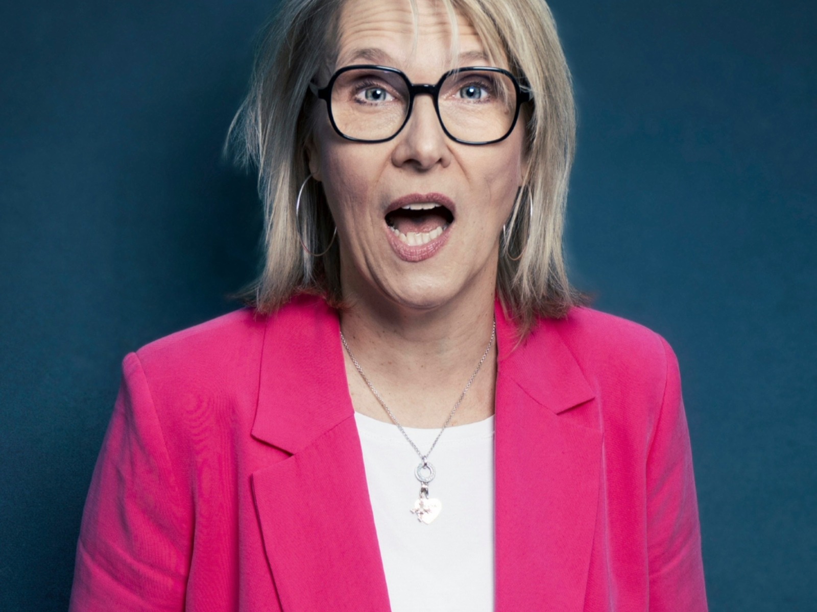 Blonde Frau mit Brille im pinken Blazer blickt überrascht mit offenem Mund vor blauem Hintergrund