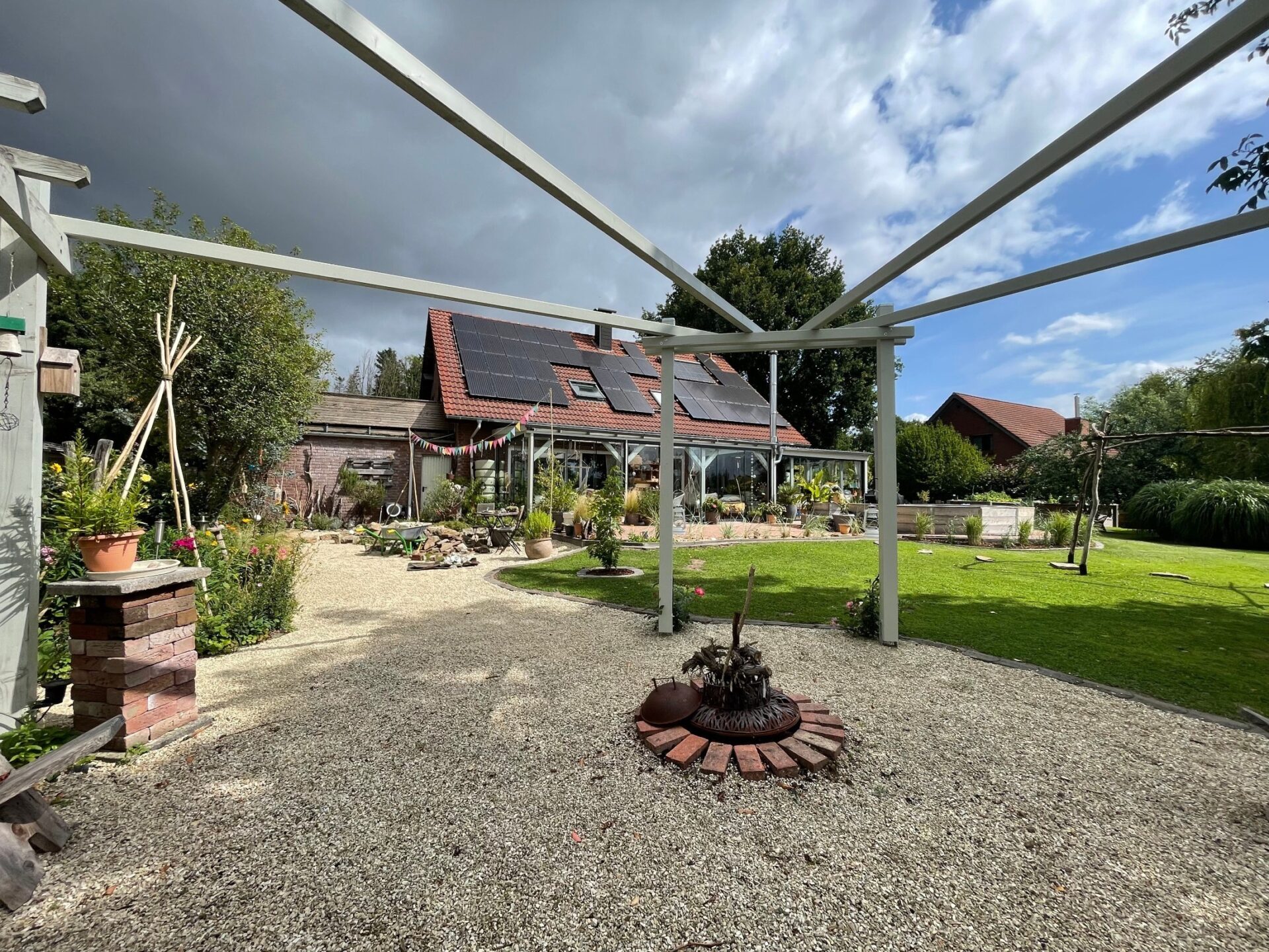 Ein gepflegter Garten mit Kiesweg, Feuerstelle, Rasen, Blumen und einem Haus mit Solardach und Wintergarten