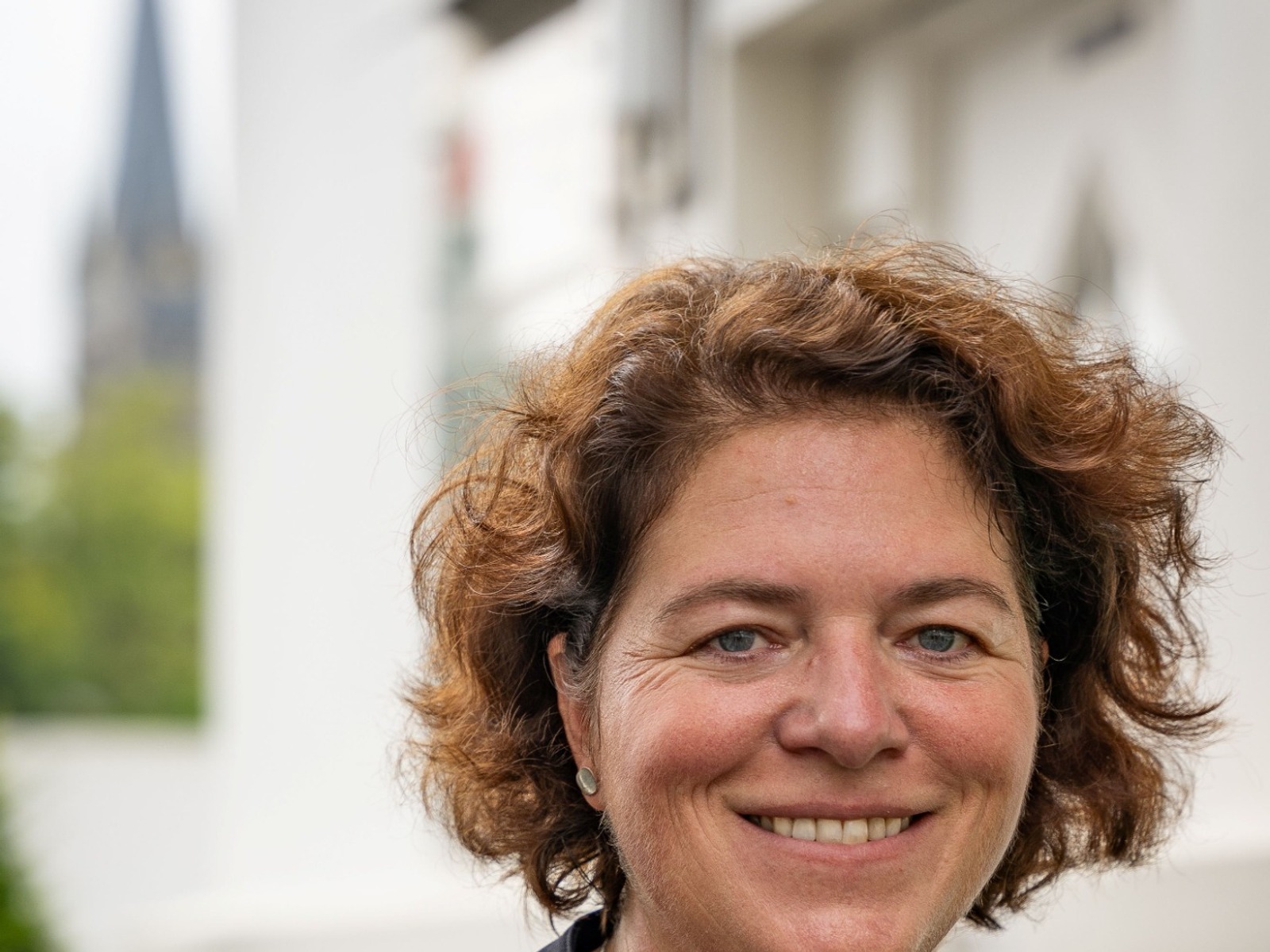 Kerstin Vieregge lächelt mit lockigen, braunen Haaren in dunkelblauer Jacke vor einer Kirche und weißem Gebäude