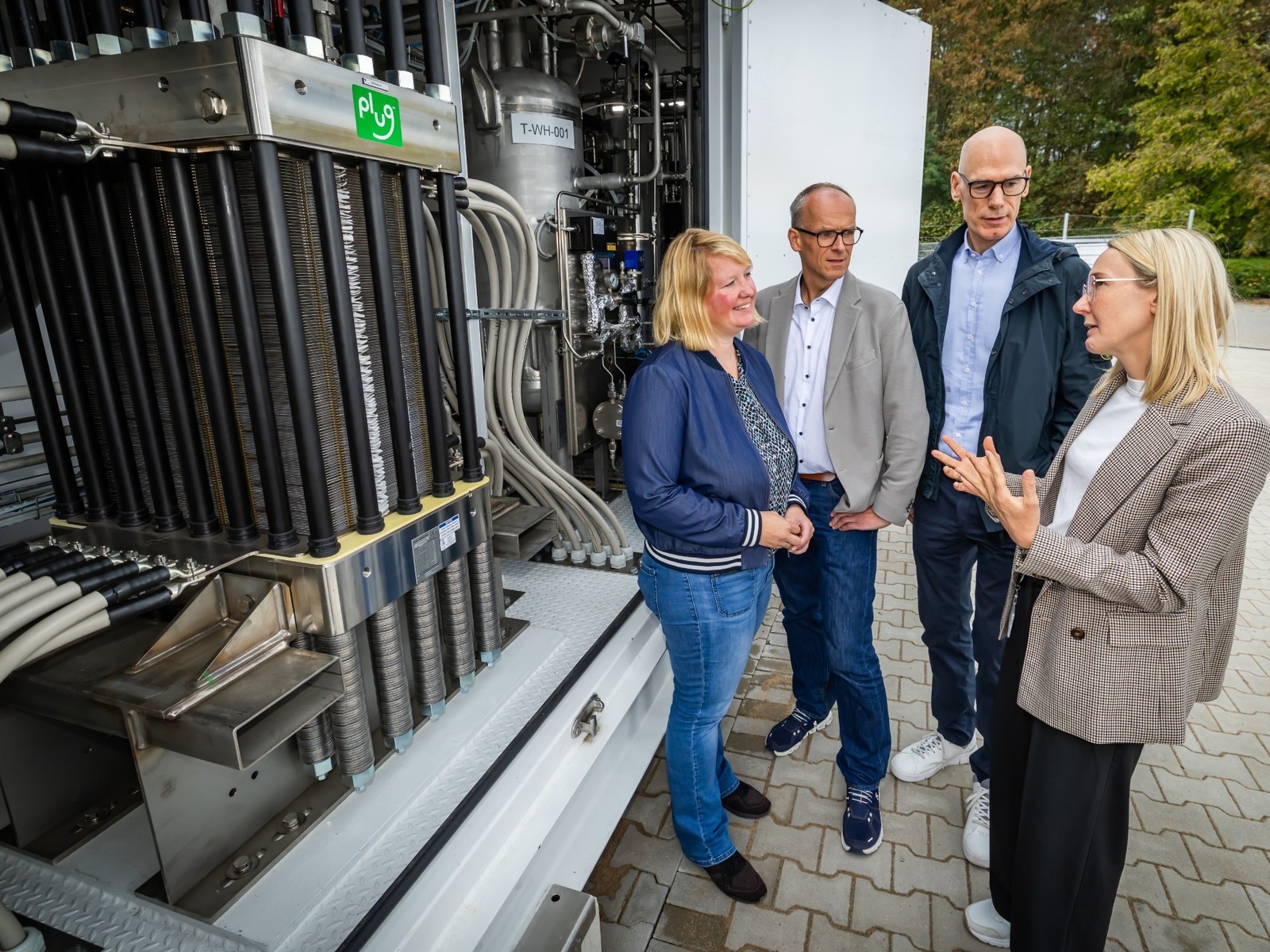 Eine Frau gestikuliert im Gespräch mit drei weiteren Personen vor einem Elektrolyseur mit Rohren und dem "Plug"-Logo