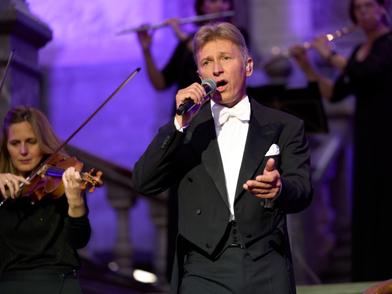 Helmut Lotti singt in schwarzem Smoking mit Mikrofon auf einer Bühne, umgeben von Musikerinnen mit Violine und Querflöte