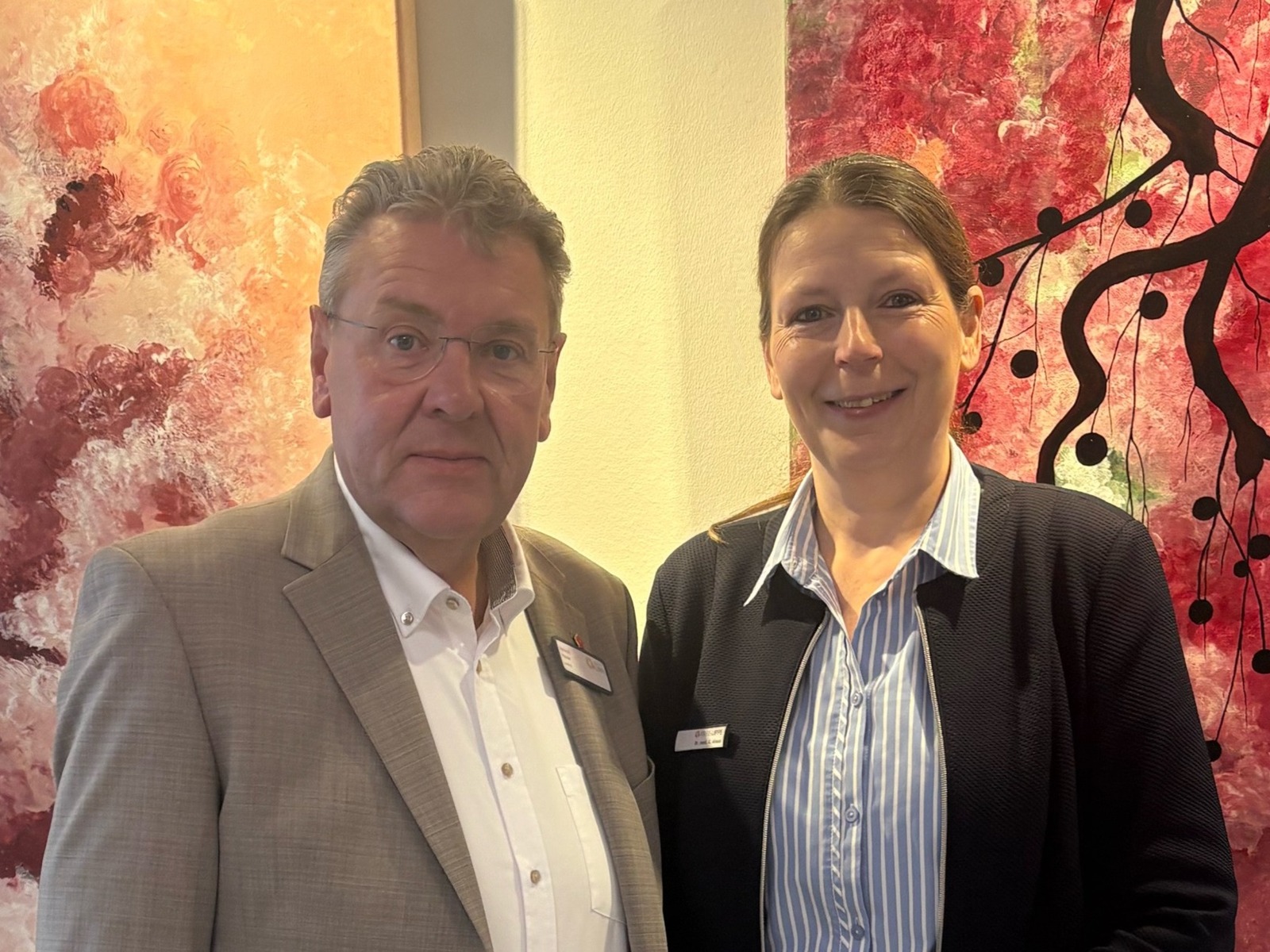 Ein Mann im Anzug und eine Frau im gestreiften Hemd mit Namensschildern stehen vor bunten Wandbildern