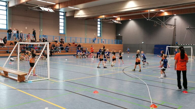 Kinder spielen Handball in einer Sporthalle mit Zuschauern auf den Tribünen und einem Erwachsenen am Spielfeldrand