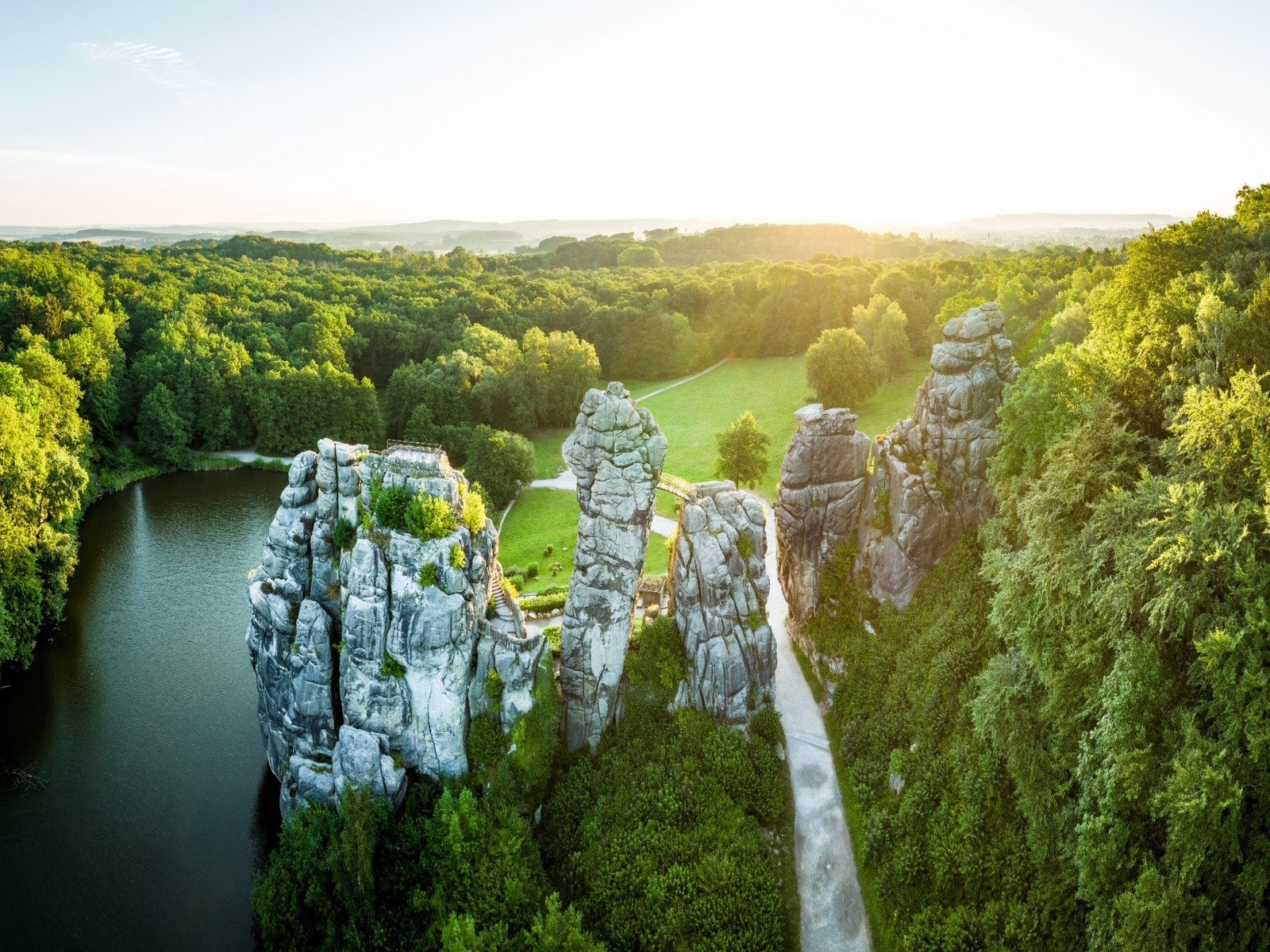 Markante Felsformation der Externsteine am dunklen See, umgeben von dichtem grünen Wald im goldenen Sonnenlicht