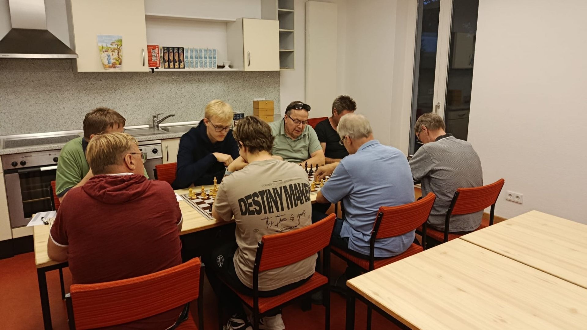 Acht Männer spielen konzentriert Schach an zwei Tischen in einem Raum mit Küchenzeile und roten Stühlen
