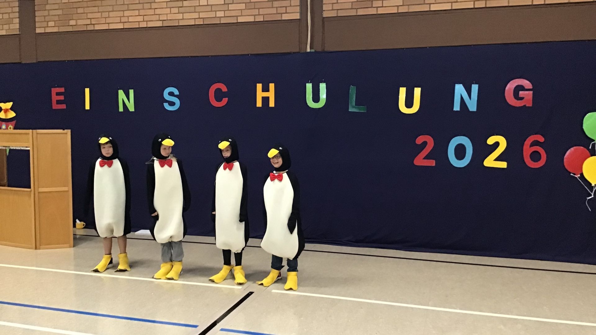 Vier Kinder in Pinguinkostümen stehen vor einem blauen Banner mit bunten Buchstaben „EINSCHULUNG 2026“ in einer Halle