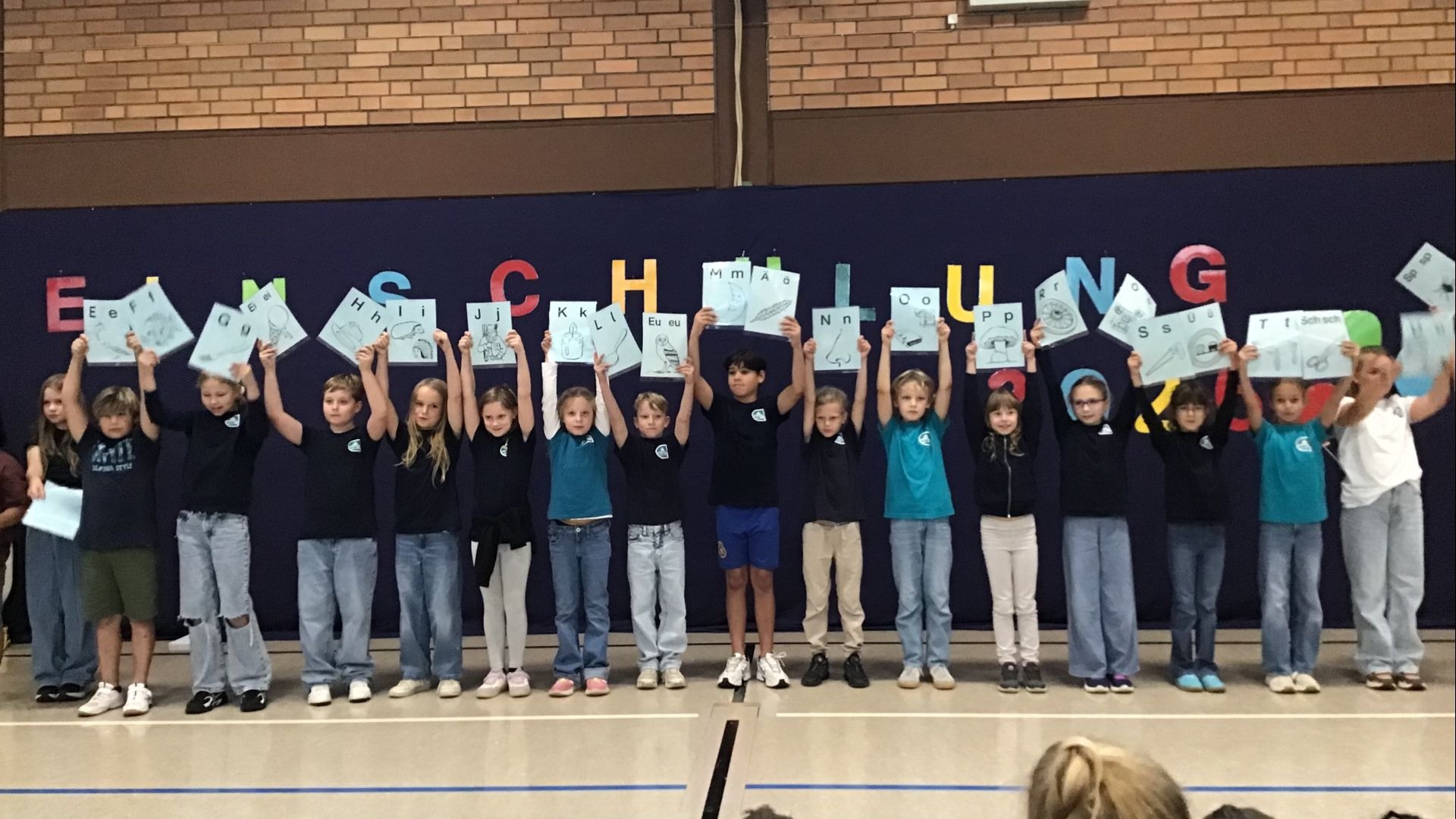 Eine Gruppe von Kindern präsentiert Blätter mit Buchstaben und Illustrationen vor einer blauen Wand mit dem Wort