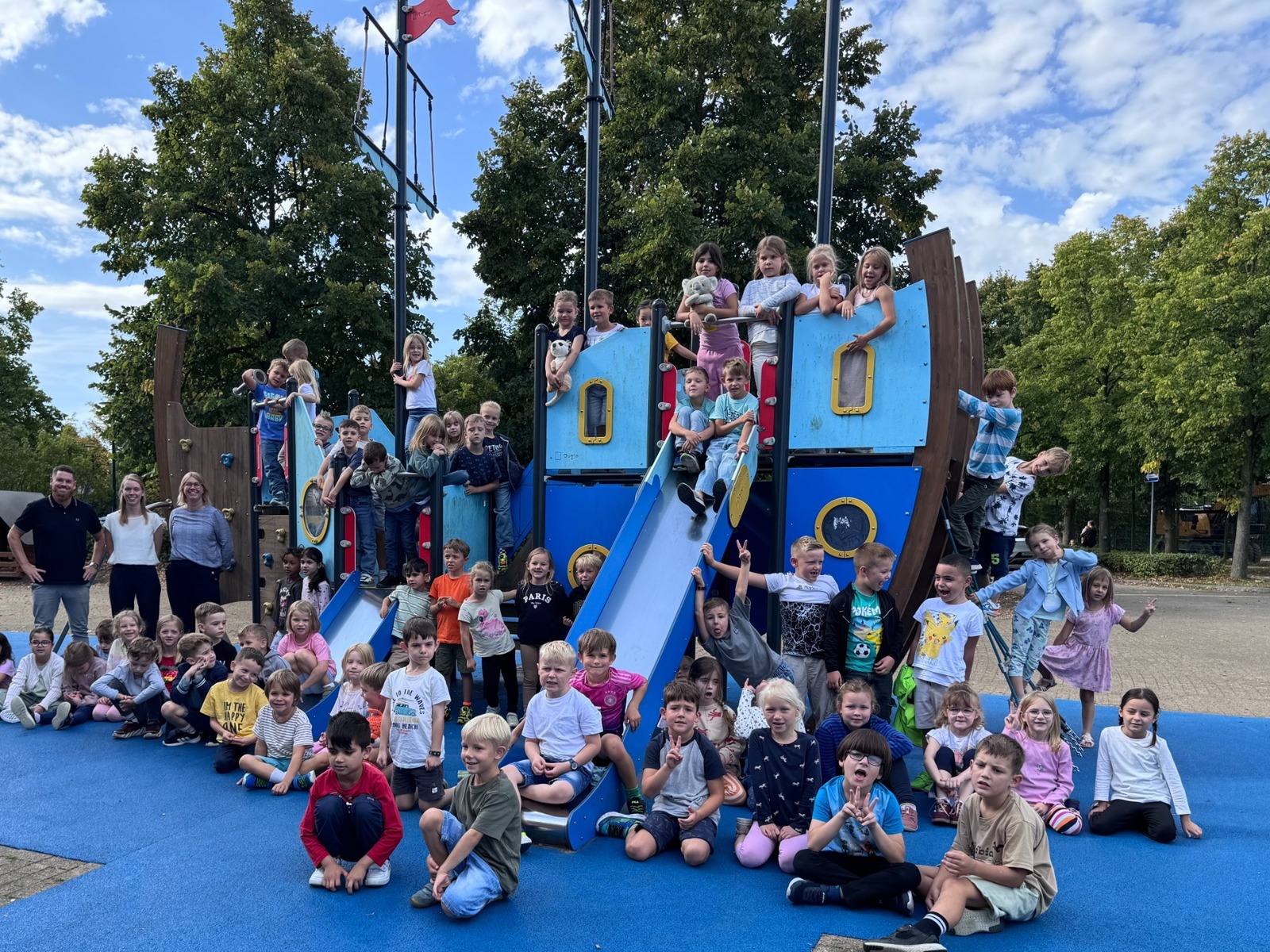 Zahlreiche lachende Kinder und einige Erwachsene auf einem blauen Spielschiff-Klettergerüst auf einem Schulhof