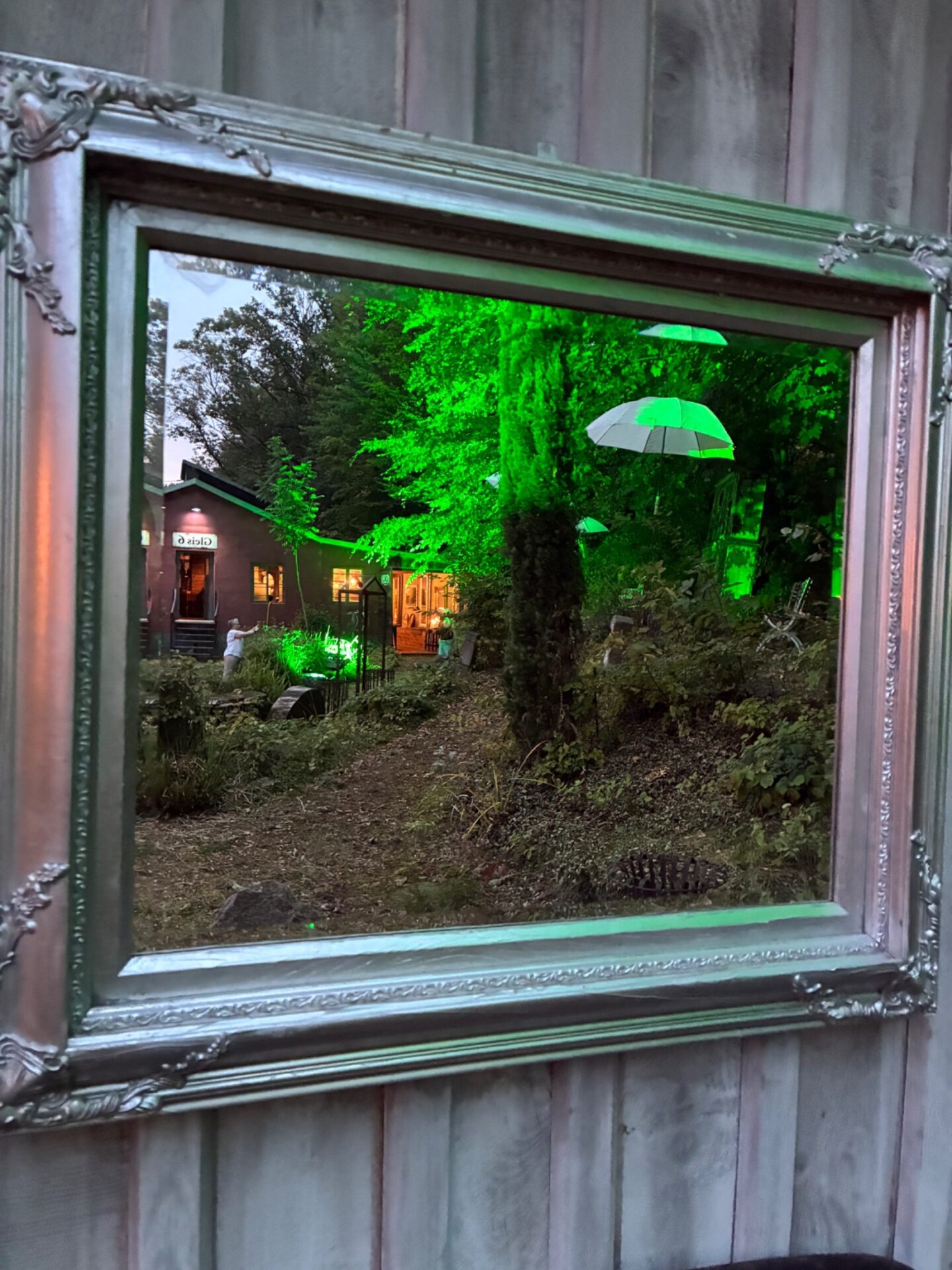 Ein antiker Silberrahmen zeigt einen nachts grün beleuchteten Garten mit hängenden weißen Schirmen, einem Gebäude und