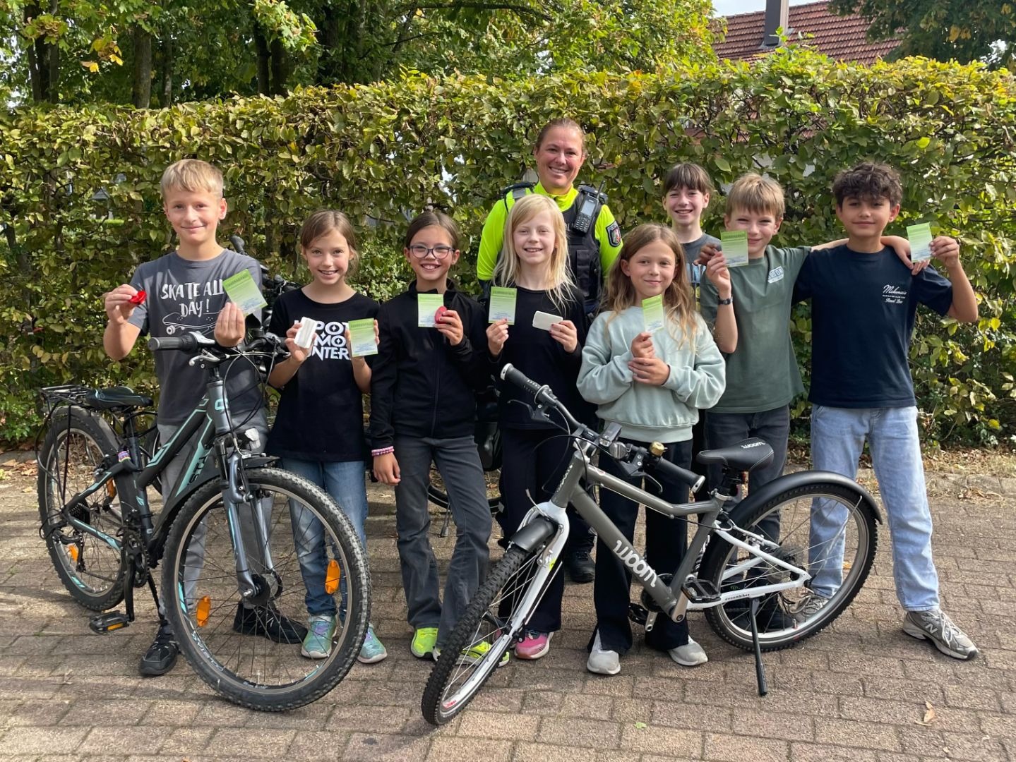 Acht lächelnde Kinder und eine Polizistin präsentieren stolz ihre Fahrradpässe neben zwei Fahrrädern vor einer grünen