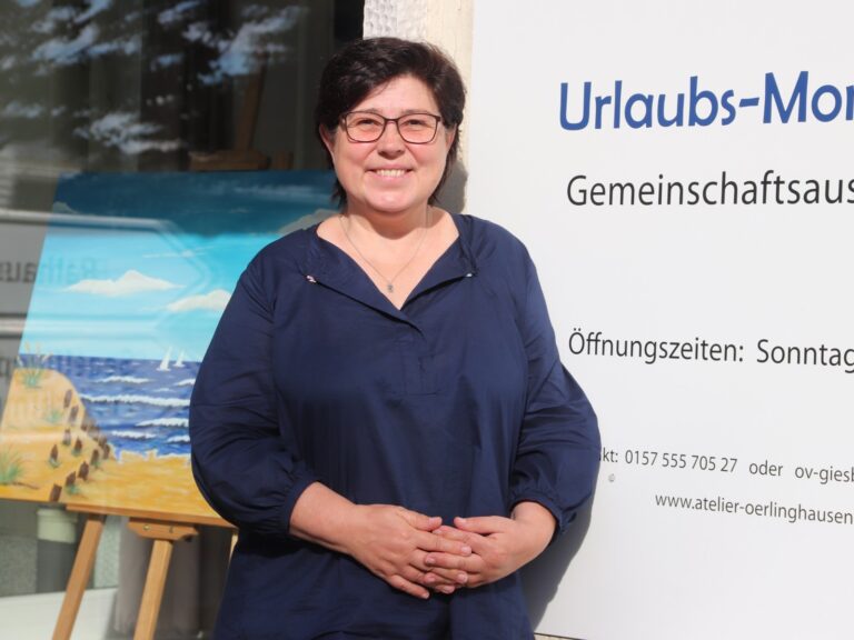 Lächelnde Frau mit Brille vor Schild "Urlaubs-Momente" und Gemälde mit Strand und Meer im Schaufenster