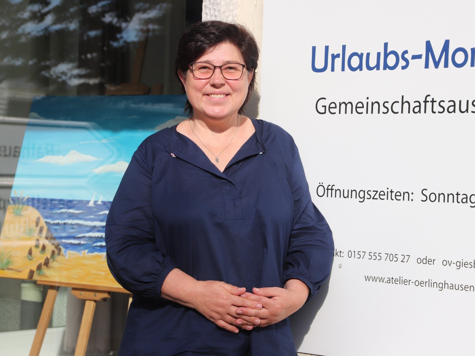 Lächelnde Frau mit Brille vor Schild "Urlaubs-Momente" und Gemälde mit Strand und Meer im Schaufenster