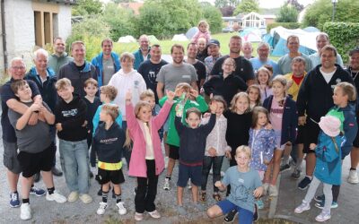 Eine große Gruppe lachender Väter und Kinder posiert für ein Gruppenfoto im Freien vor aufgebauten Zelten und einem