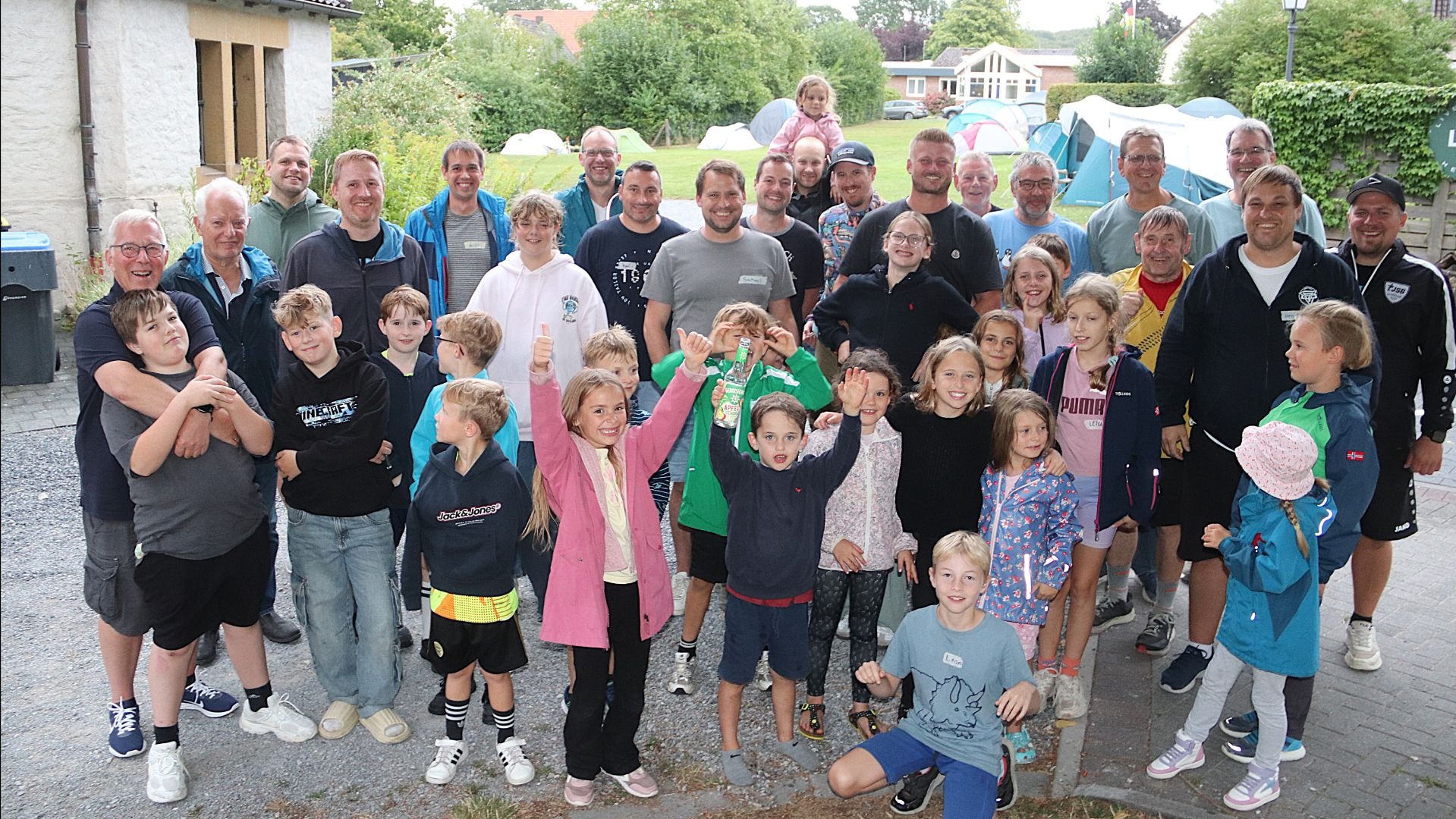 Eine große Gruppe lachender Väter und Kinder posiert für ein Gruppenfoto im Freien vor aufgebauten Zelten und einem