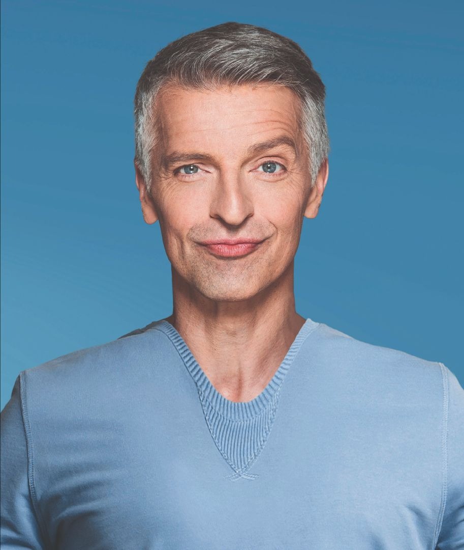 Johannes Flöck lächelt leicht, trägt ein hellblaues Shirt und hat grau meliertes Haar vor blauem Hintergrund