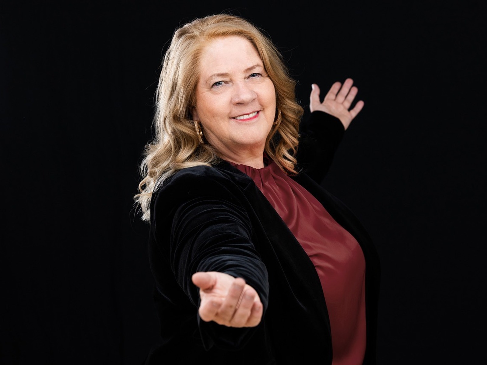Kathy Kelly lächelt und macht eine einladende Geste mit beiden Armen zum Betrachter