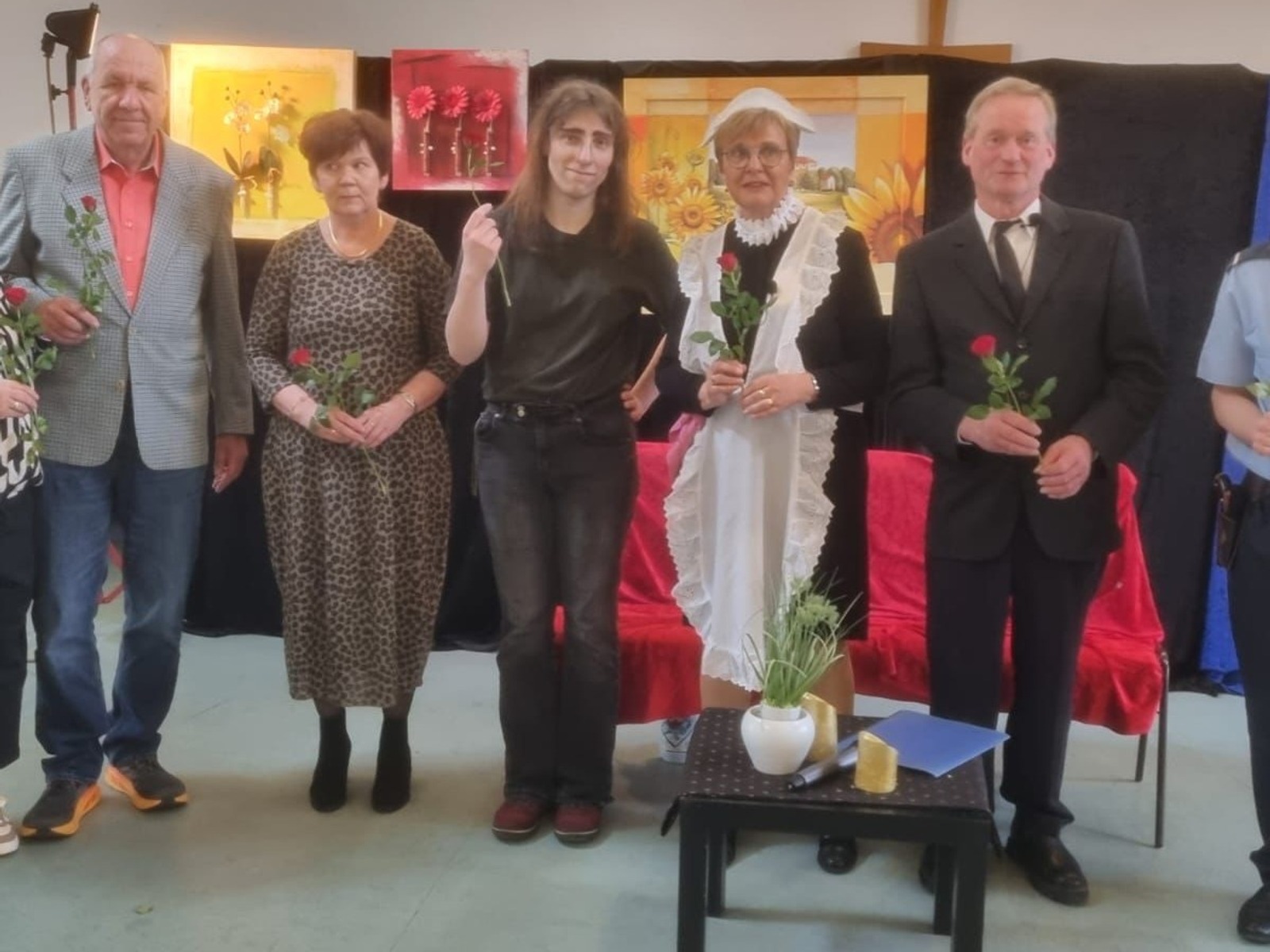 Sechs Schauspieler einer Laienspielgruppe, teils in Theaterkostümen, halten rote Rosen auf einer Bühne