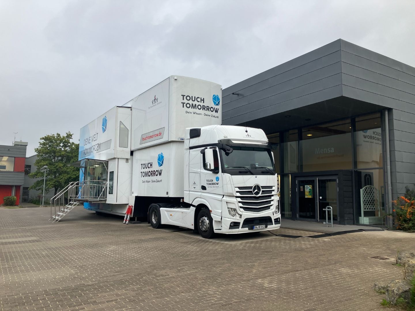 Weißer TouchTomorrow-Truck mit ausgefahrenem Aufbau und Treppe parkt vor einem Gebäude mit „Mensa“-Beschriftung