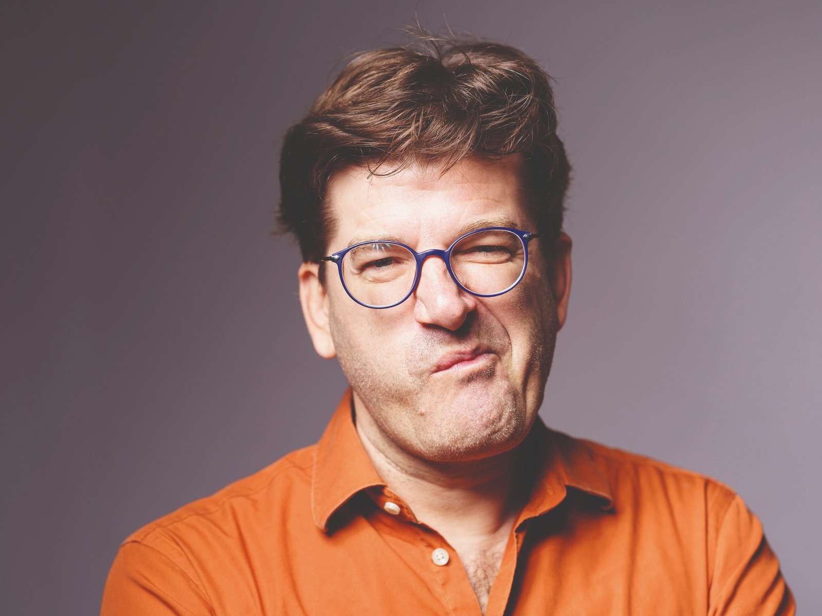 Nils Heinrich mit blauer Brille und orangefarbenem Hemd grimassiert mit verzogenem Mund und zusammengekniffenen Augen