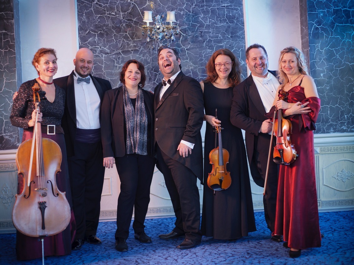 Drei Männer und drei Frauen in festlicher Abendkleidung lächeln mit Cello und zwei Geigen in einem Saal