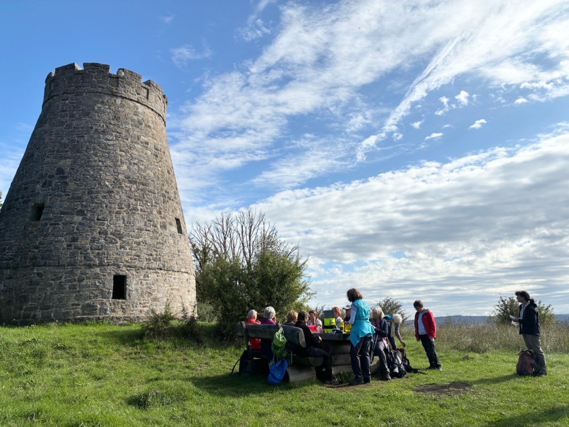 Wandergruppe macht Pause an einem Holztisch neben einem alten Steinturm auf einer grünen Wiese unter blauem Himmel