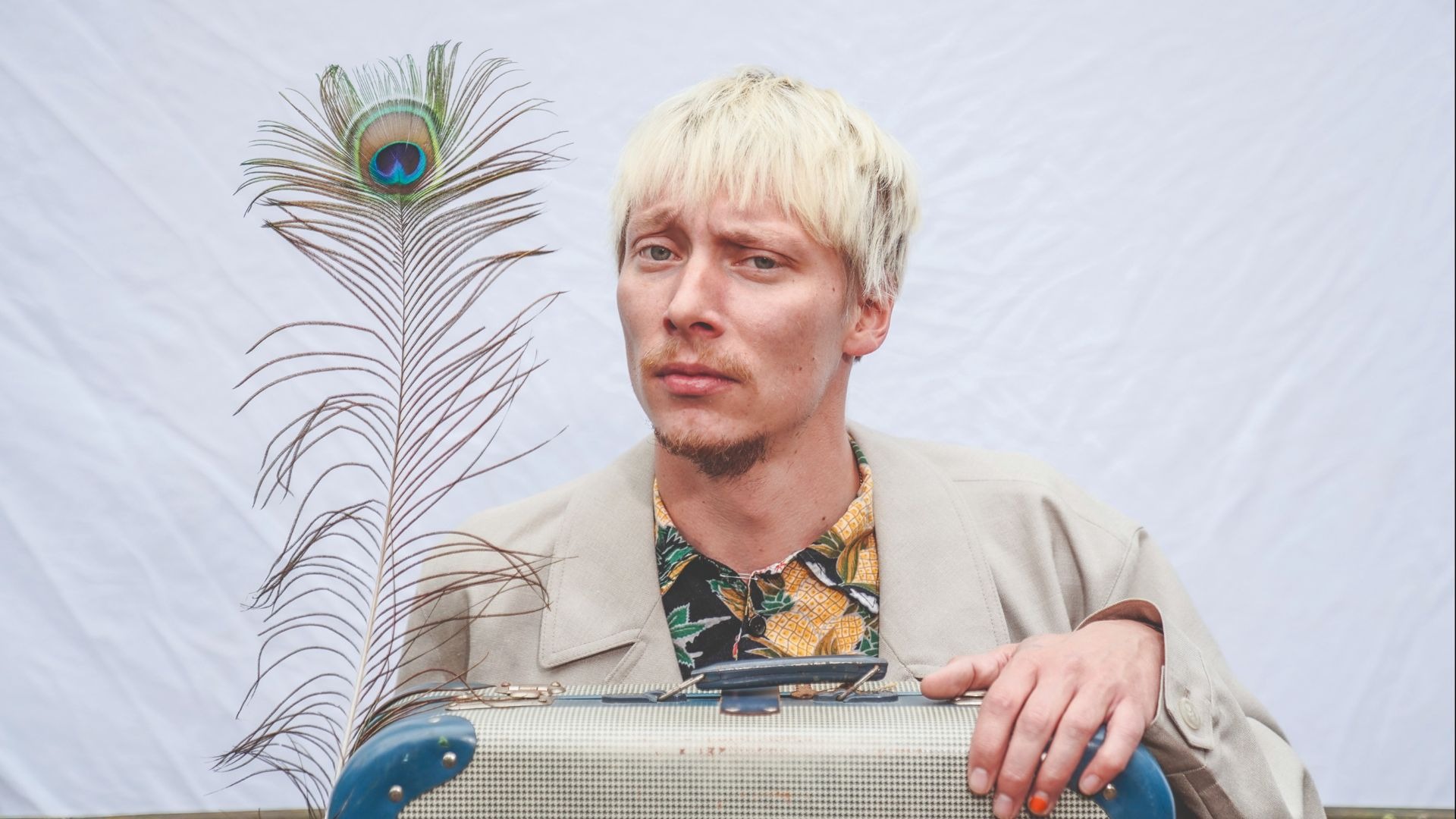 Robert Alan mit blonden Haaren und Bart in Jacke und Hemd, stützt sich auf einen Koffer, daneben eine Pfauenfeder