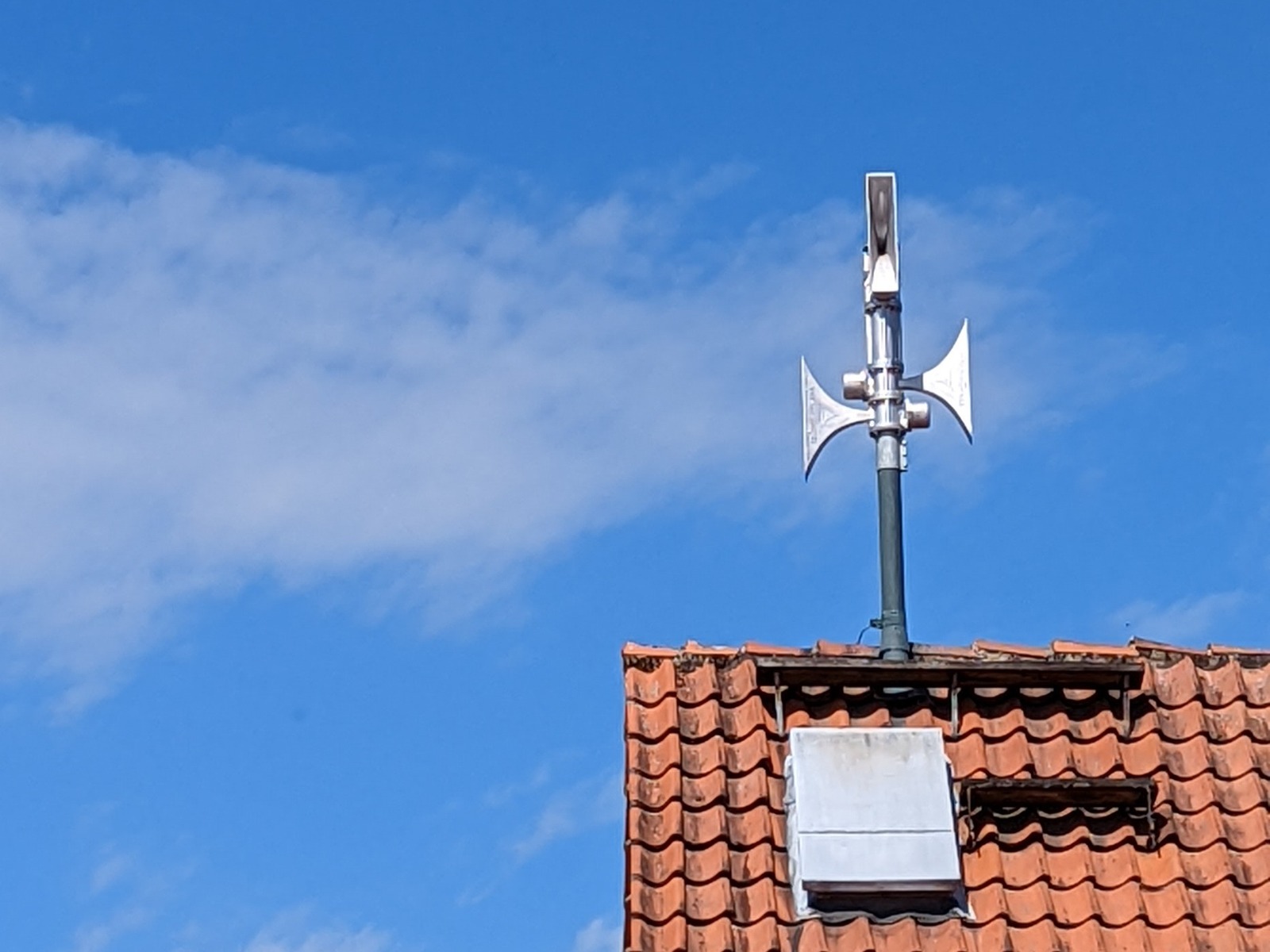 Eine metallene Sirene mit mehreren Lautsprechern auf einem roten Ziegeldach unter blauem Himmel