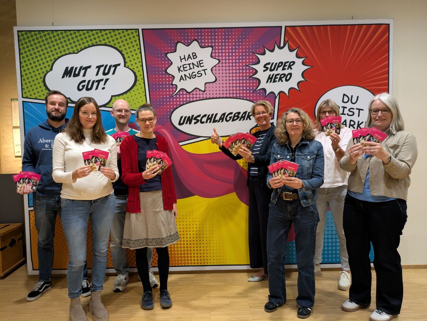 Acht lächelnde Personen präsentieren rosa Programmhefte vor einer Comic-Wand mit Sprechblasen und Motivationssprüchen