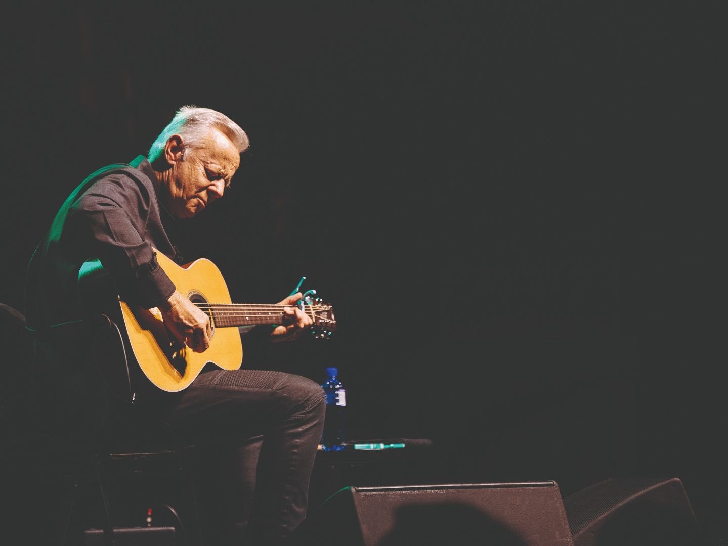 Tommy Emmanuel beugt sich über seine Akustikgitarre und spielt auf einer dunklen Bühne im Scheinwerferlicht