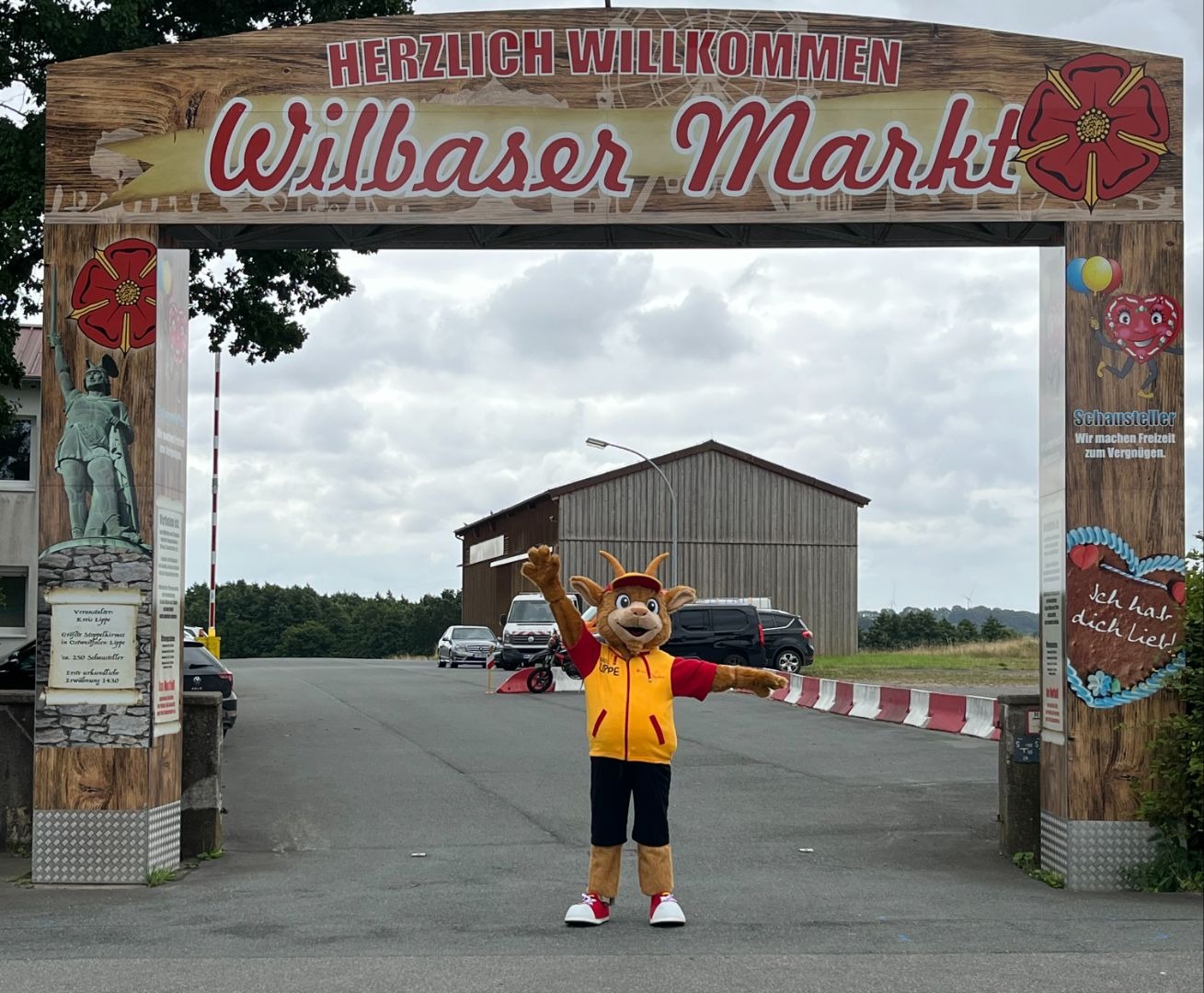 Wilbaser Markt Eingangsportal mit Maskottchen Lippi in gelb-roter Kleidung, das die Arme ausbreitet auf Asphaltfläche