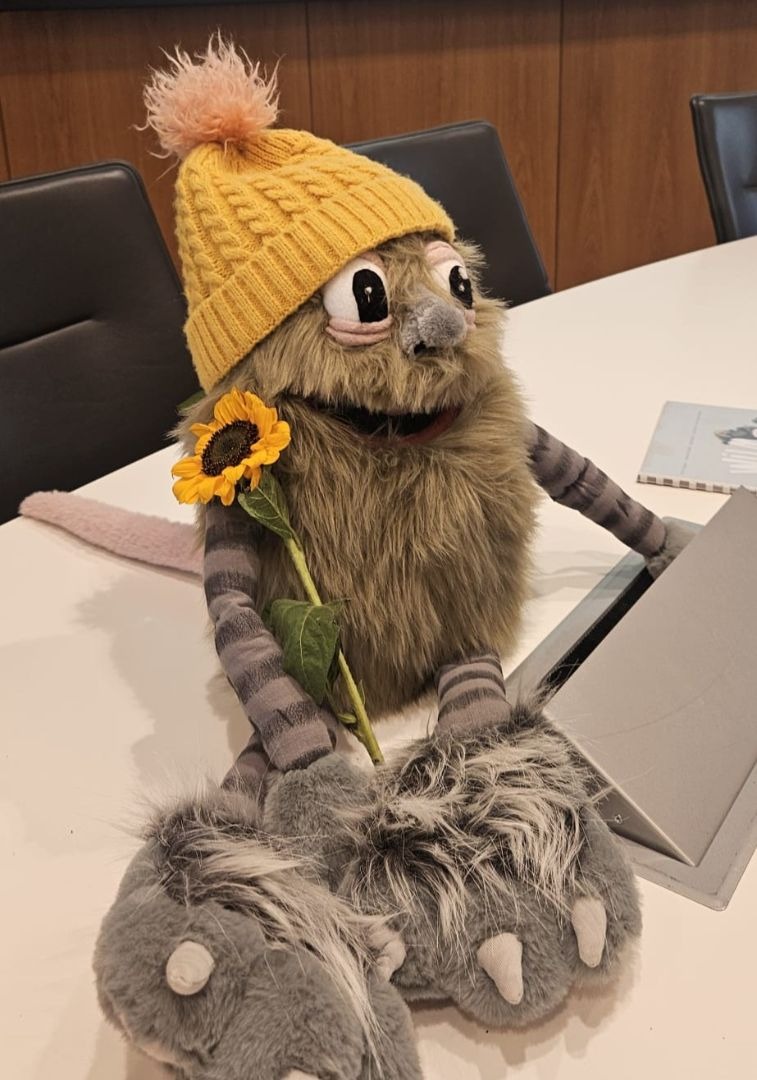 Ein felliges Stofftier mit gelber Strickmütze und Sonnenblume sitzt auf einem Tisch vor einer Präsentationsfolie mit
