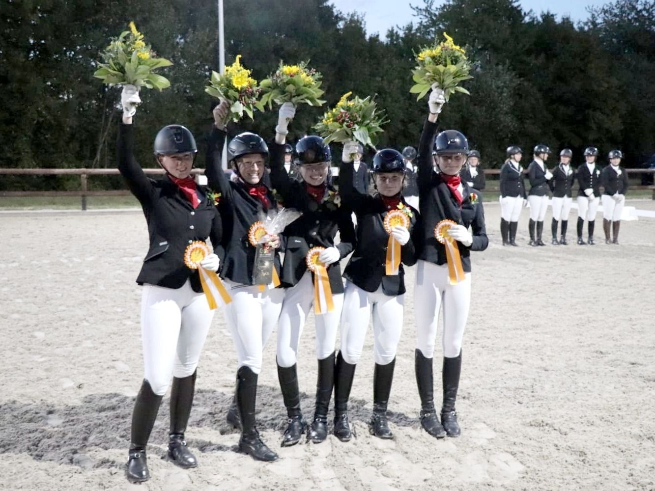 Fünf lächelnde Reiterinnen in Uniform halten Blumensträuße und Siegerschleifen in einer Sandreitbahn hoch