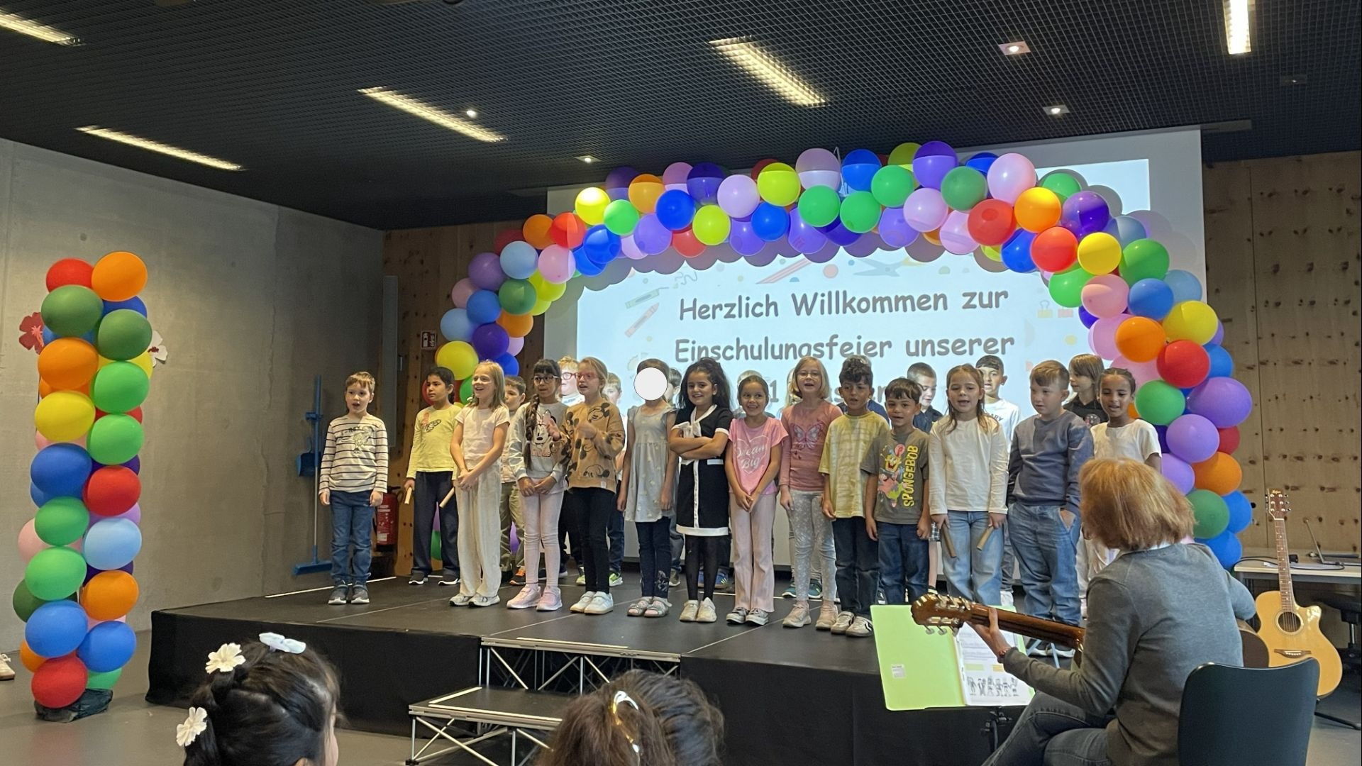 Kinder stehen auf einer Bühne vor einem bunten Ballonbogen und einer Projektion mit "Einschulungsfeier", eine Frau