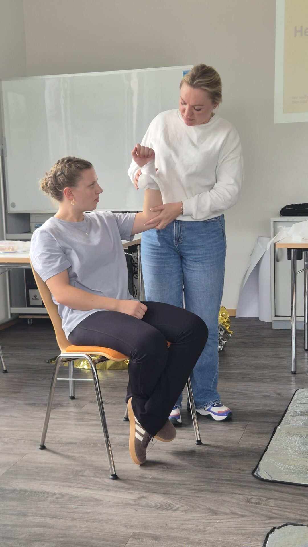Eine stehende Frau legt im Erste-Hilfe-Kurs einer sitzenden Frau einen Verband um den Unterarm