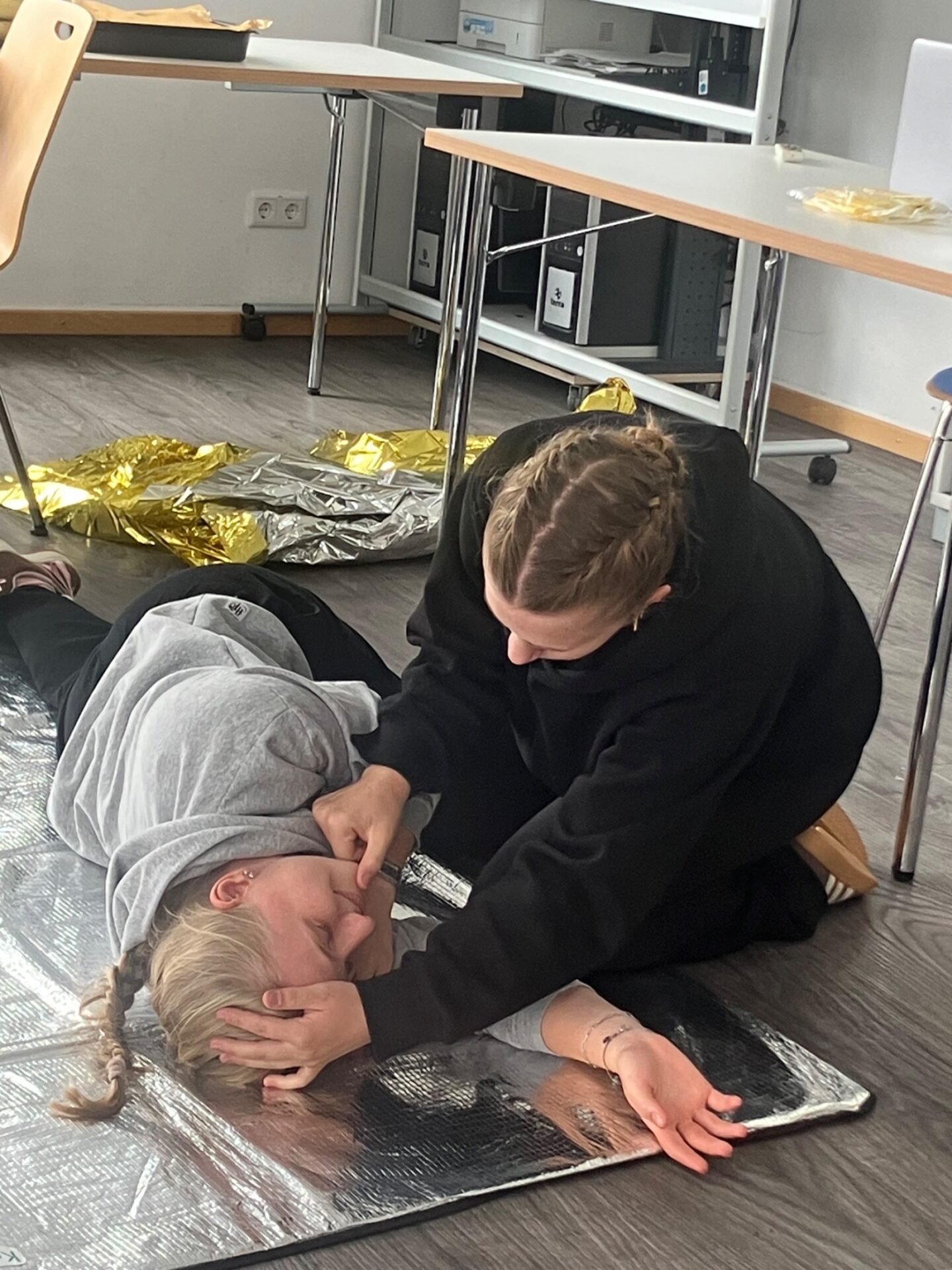 Frau kniet und überprüft die Atemwege einer auf Rettungsdecke liegenden Person während eines Erste-Hilfe-Kurses