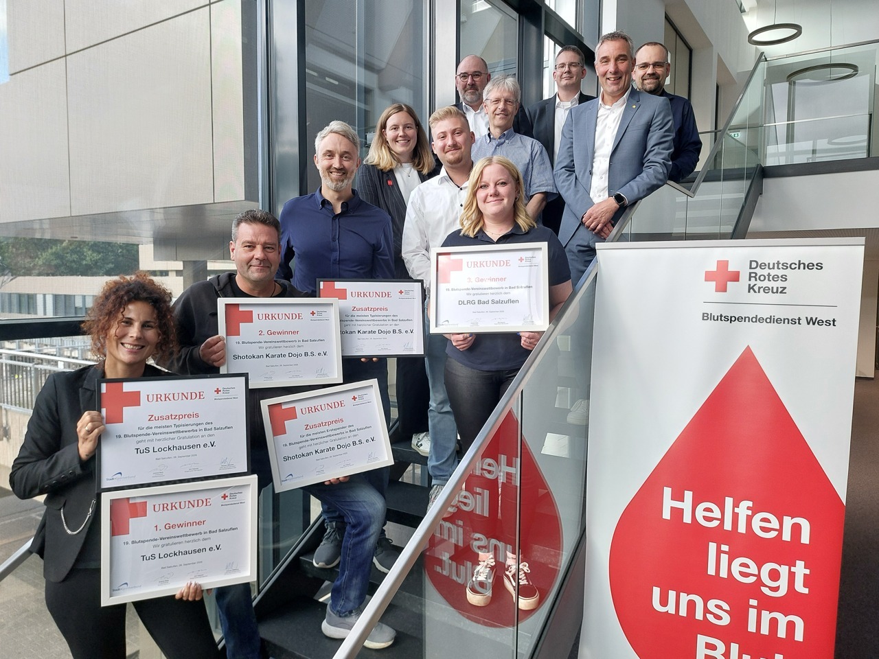 Ein Dutzend Männer und Frauen lächeln auf einer Treppe und halten DRK-Urkunden für Blutspende-Wettbewerbe