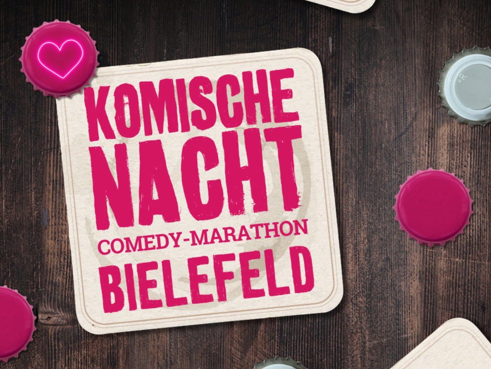 Ein Bierdeckel mit der Aufschrift 'Komische Nacht Comedy-Marathon Bielefeld' liegt auf dunklem Holz umgeben von